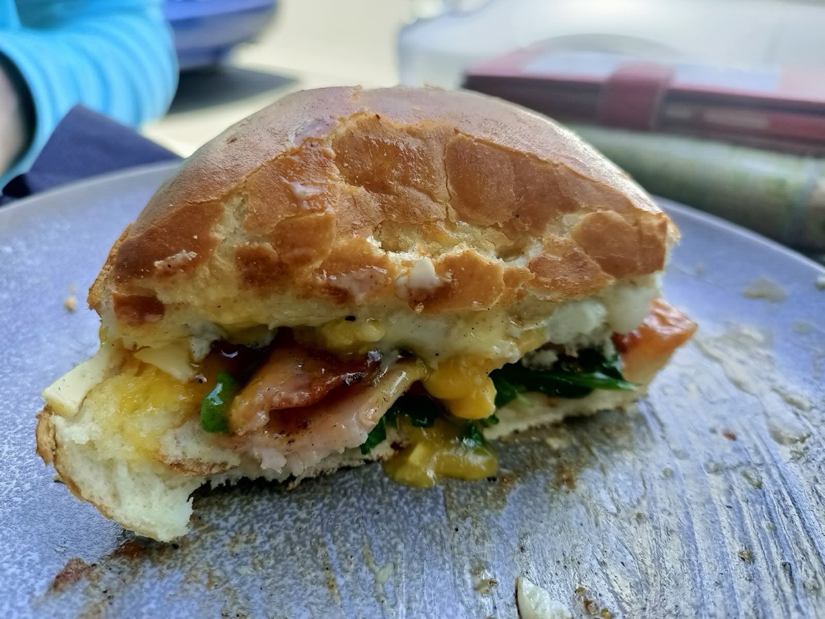 A half-eaten toasted bun filled with egg, bacon, greens and cheese, on a blue-grey plate