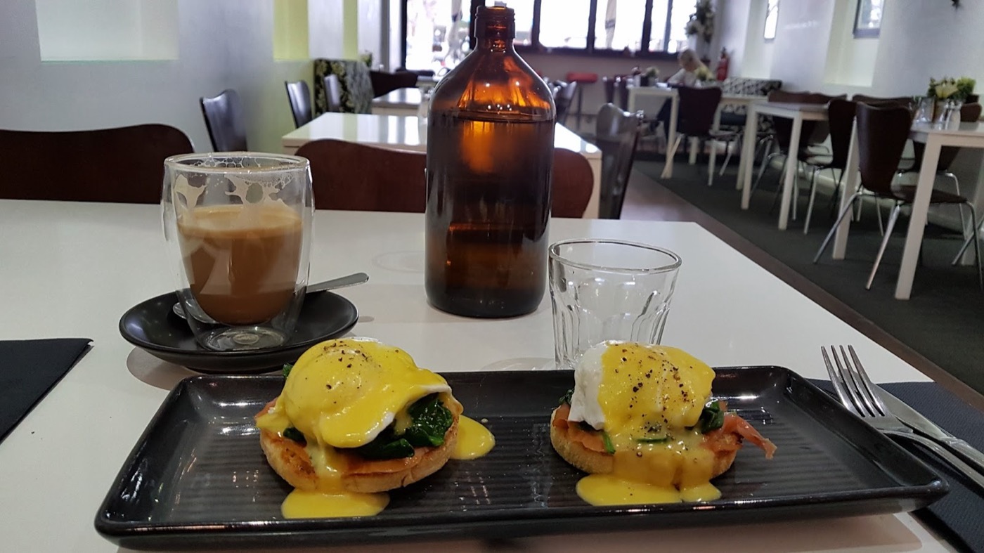 A black tray with two muffin stacks topped with poached egg, greens and yellow hollandaise, beside an iced coffee and a brown bottle