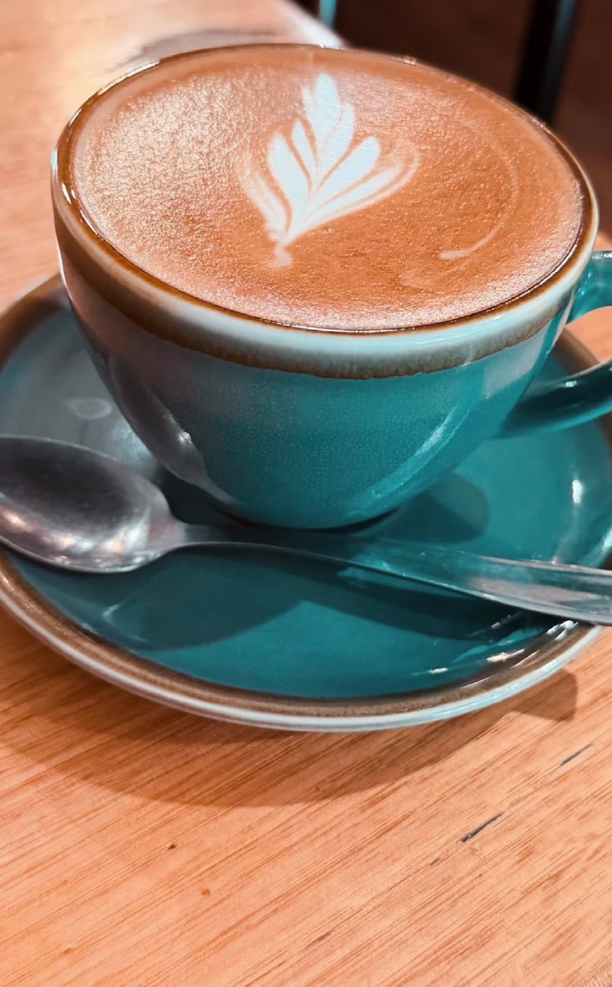 A latte with a leaf pattern in the foam, in a teal cup on a teal saucer, on a pale timber table