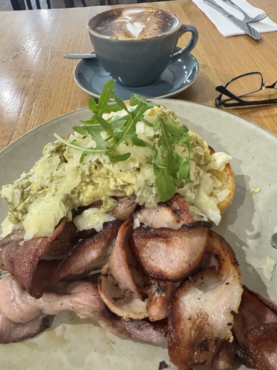 Scrambled eggs piled on thick-cut bacon on toast, with rocket on top and a coffee cup behind